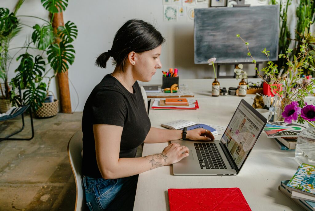 A woman sits at a desk in a creative space, using a laptop surrounded by plants and art supplies.